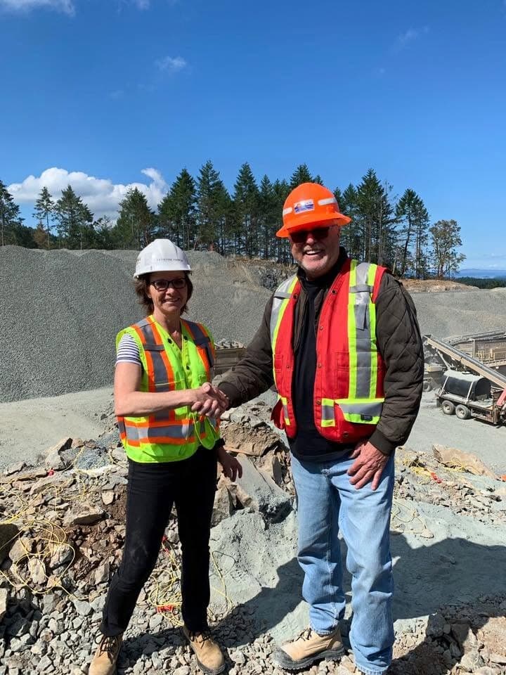 Katherine Pankiw shaking hands with a colleague in hi-vis gear at the Cantex Summerland pit, crushed-rock stockpiles and a crusher behind them.